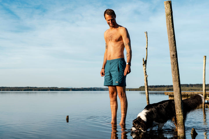 Man standing in shallow lake water in teal shorts beside his collie