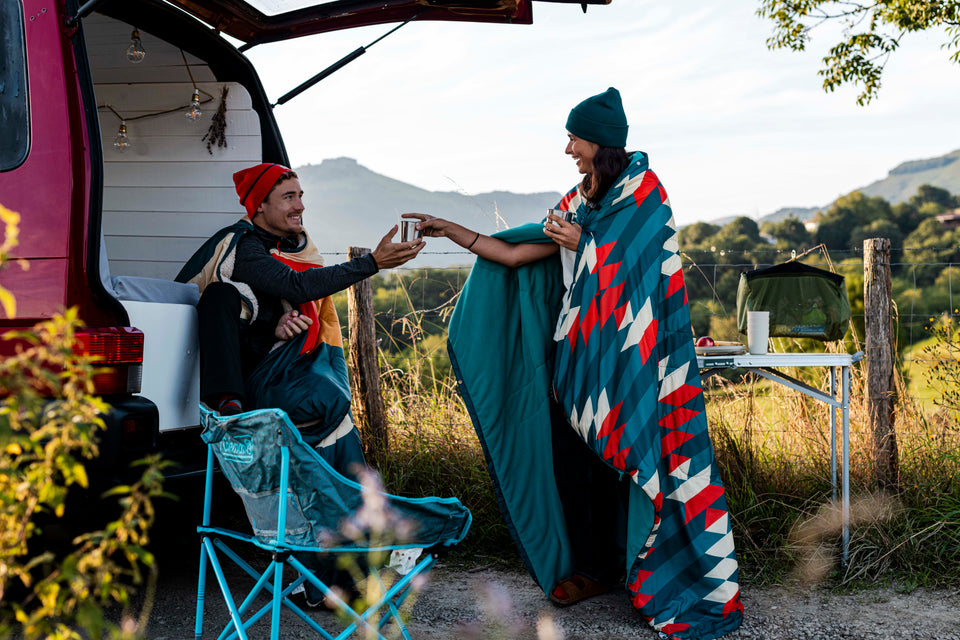 ouple wrapped in patterned blankets sharing coffee beside a camper van