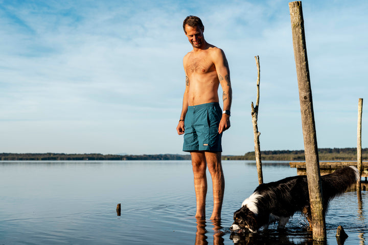Man standing in shallow lake water in teal shorts beside his collie