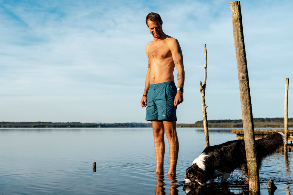Man standing in shallow lake water in teal shorts beside his collie