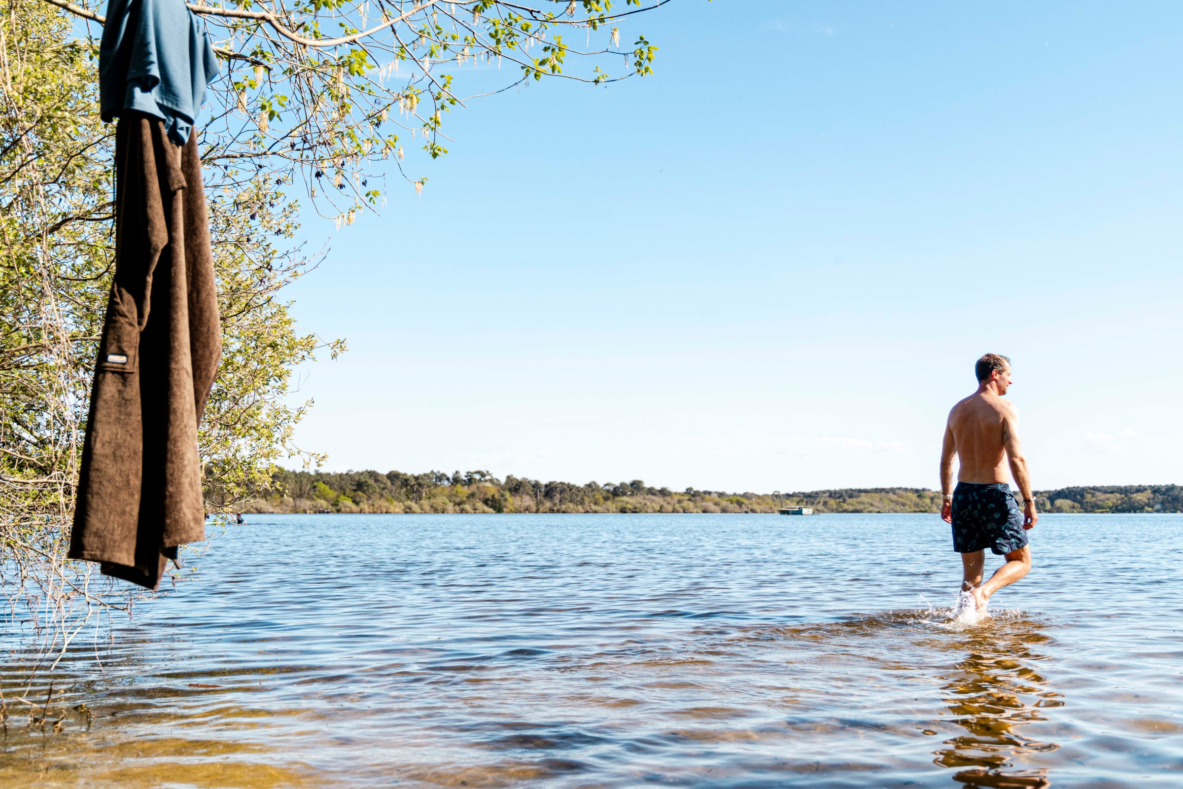 Man wading into a calm lake in patterned shorts, towel and tee drying on a branch