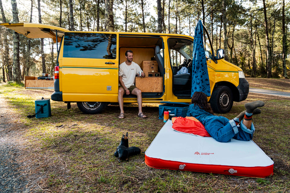 A yellow camper van parked in the woods with a man sitting by the open side door and a woman lying on a red and white camping mattress on the ground