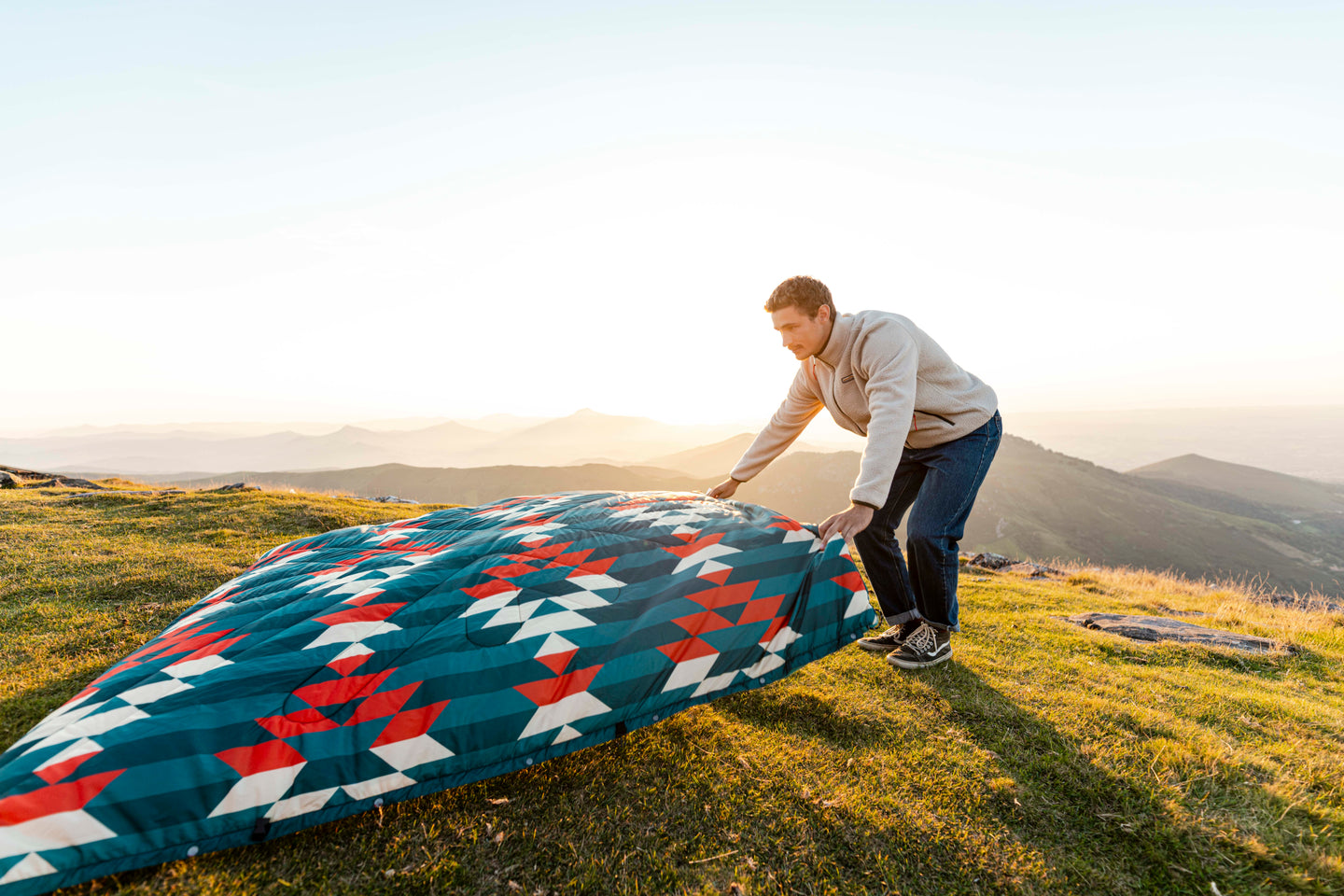 Man laying out a sherpa blanket on a hilltop at sunset