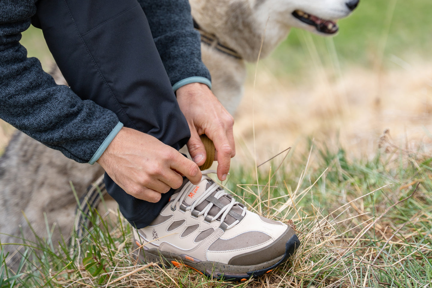 Person tying beige hiking shoes beside a dog in a grassy field