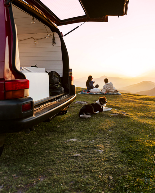 Two people and a dog sitting on a blanket in front of an open van