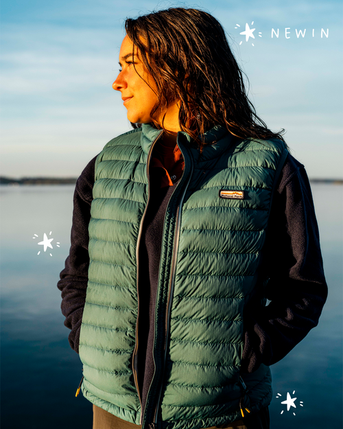 A woman wearing an insulated gilet over a dark long-sleeve fleece, standing outdoors by a calm lake at sunset