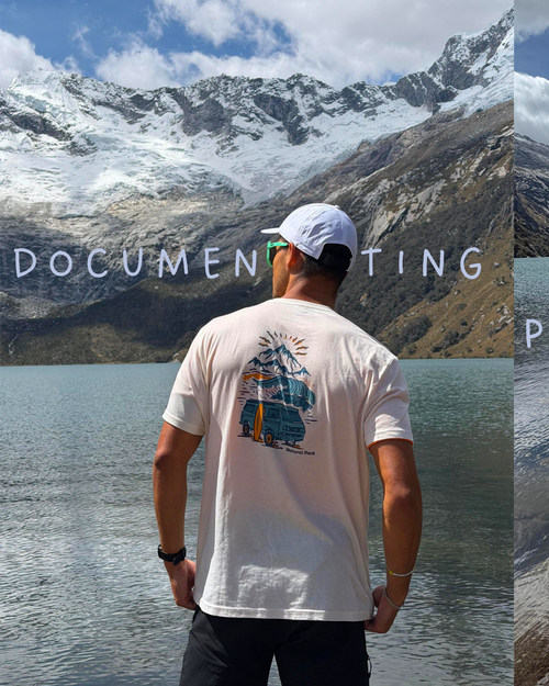 A man stands with his back to the camera in front of a mountain and lake, wearing a white graphic t-shirt