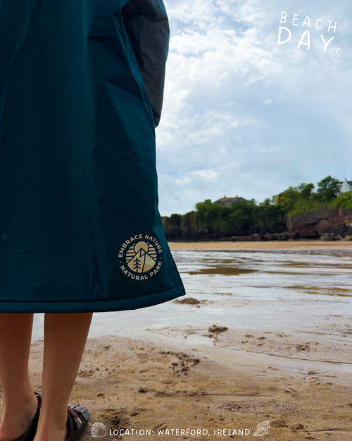 A person wearing a dark teal waterproof changing robe stands on a sandy beach