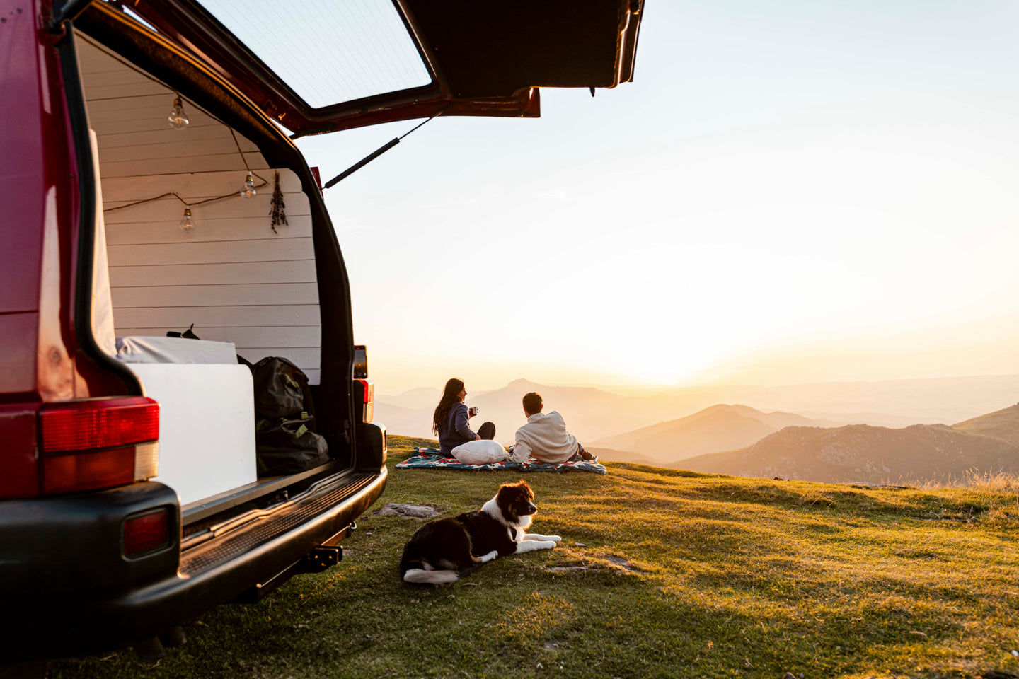 Couple sitting beside a camper van at sunset with a dog on a blanket