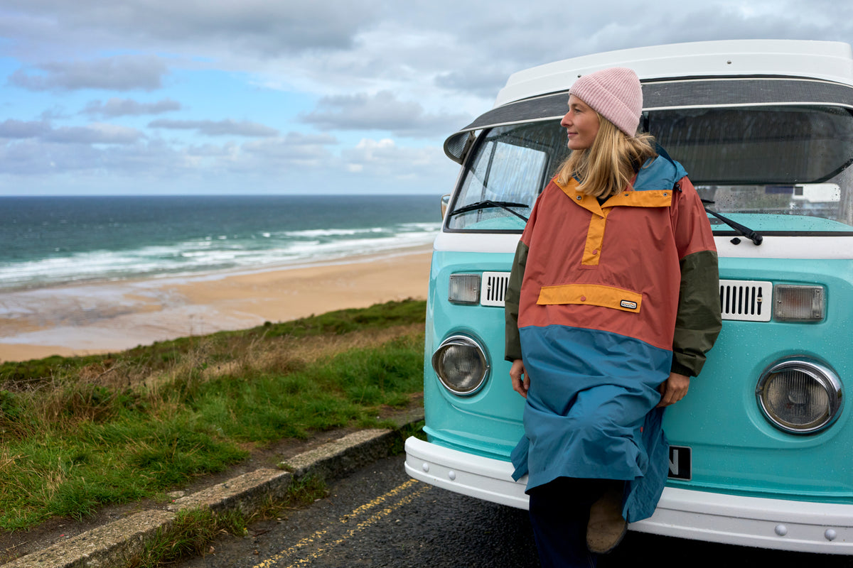 A person in a waterproof poncho leaning against a retro campervan at the seaside