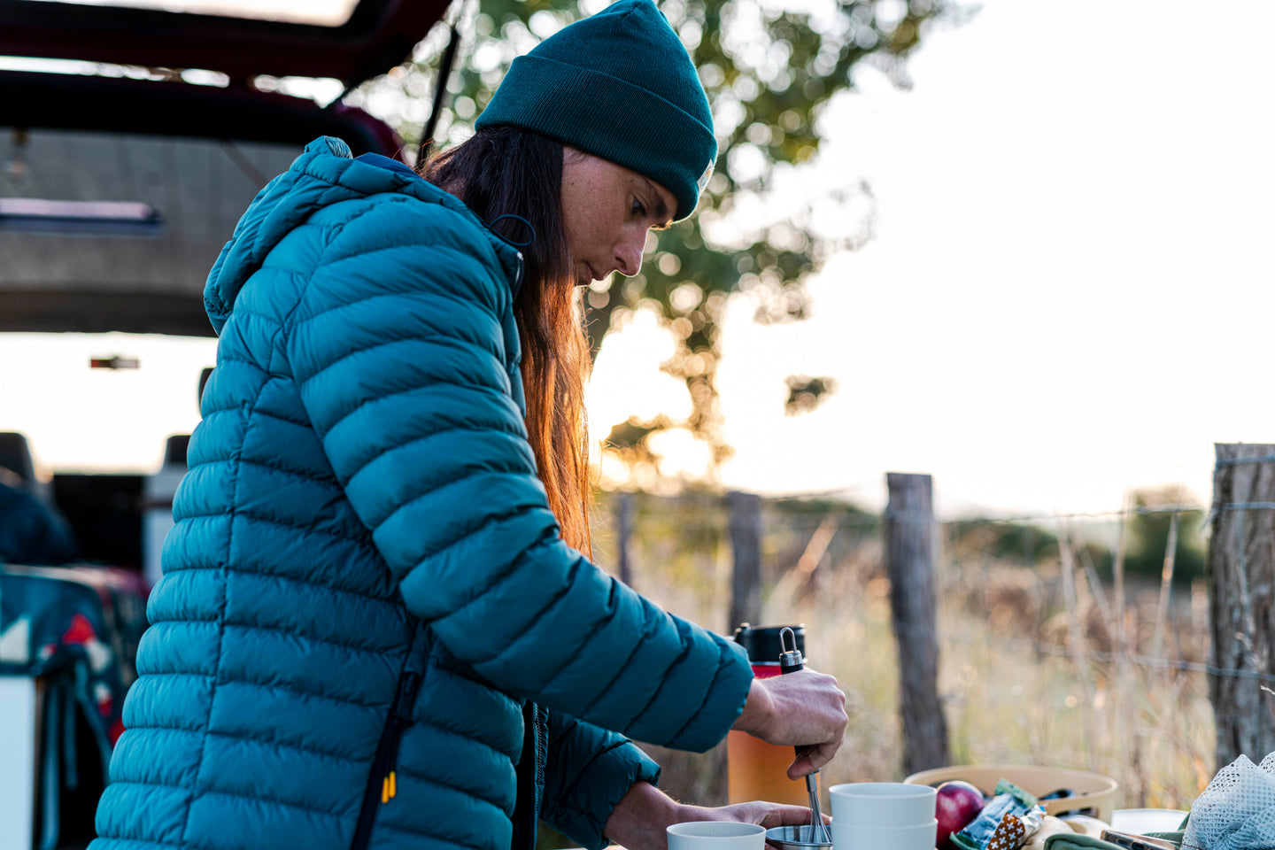 Woman wearing a teal insulated jacket and beanie making coffee